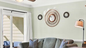 A living room with a grey sofa, striped pillows, a wooden coffee table, and a dining table. There are two wall-mounted circular mirrors and a floor lamp. - Great Beach Condo, Across From Heated Pool Access (Tybee Island)