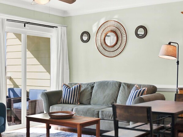 A living room with a grey sofa, striped pillows, a wooden coffee table, and a dining table. There are two wall-mounted circular mirrors and a floor lamp. - Great Beach Condo, Across From Heated Pool Access (Tybee Island)