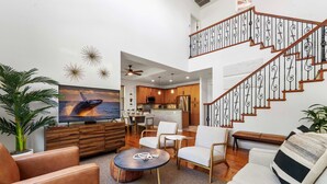 A spacious living area with a large TV, comfortable seating, a staircase, and a kitchen in the background. - 🏖️Private Lanai | Golf/Sunset Views | Pool | Keyless (Kamuela)