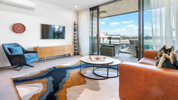 A modern living room with a large window, a flat-screen TV, a blue armchair, a brown leather sofa, and a patterned rug.