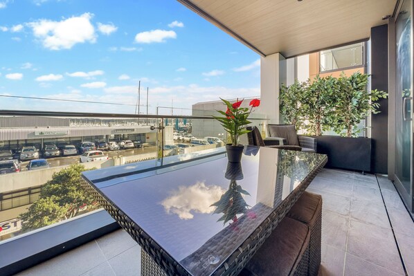 A balcony with a glass table, potted plants, and a view of a parking lot and buildings. - Beautiful and bold 1 bed retreat (Auckland)