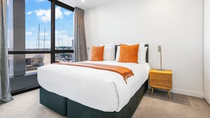A modern hotel room with a large bed, a nightstand, and a view of the city through large windows.