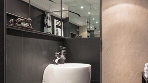 A modern bathroom with a large white sink, dark tiled walls, and a mirror reflecting the room.
