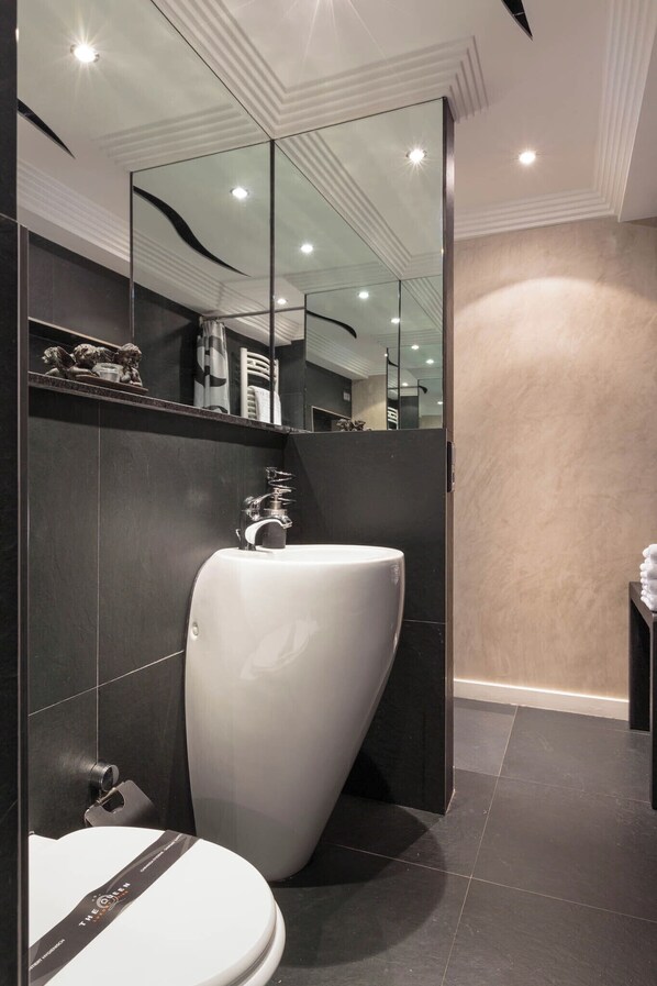 Un baño moderno con un lavamanos grande de color blanco, paredes cubiertas de azulejos oscuros y un espejo que refleja la habitación.