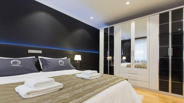 A modern bedroom with a large bed, a wardrobe with mirrored doors, and a bedside table with a lamp.