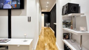 A modern hallway with a wooden floor, a TV mounted on the wall, a white desk with a lamp, and a shelf with books and a speaker.