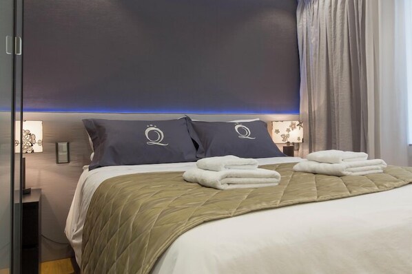Una cama bien tendida con un edredón verde acolchado, dos almohadas con un logotipo circular y toallas blancas dobladas.