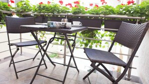 Un balcón con una mesa y dos sillas, rodeado de macetas con flores rojas.