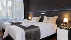 Ein Hotelzimmer mit zwei Betten, einem dunklen, strukturierten Kopfteil, einem Nachttisch mit Lampe und einem Fenster mit durchsichtigen Vorhängen.