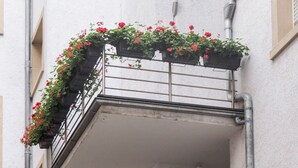Ein mehrstöckiger Balkon mit Blumenkästen, in denen rote Blumen blühen, einem Metallgeländer und einem Tisch mit Stühlen.