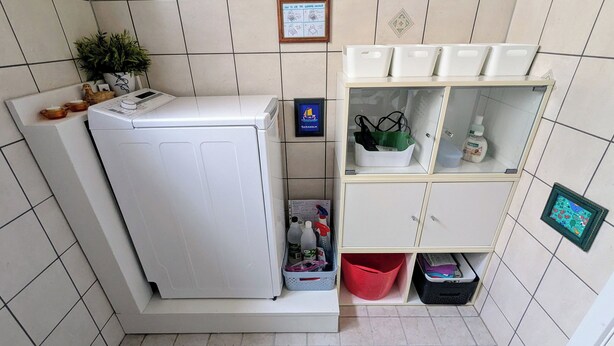 Ett kompakt kök med tvättmaskin, en liten växt, en hylla med förvaringslådor och en inramad tavla på väggen.