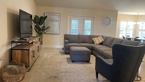 Een woonkamer met een hoekbank, voetenbankje, televisie en een grote plant.