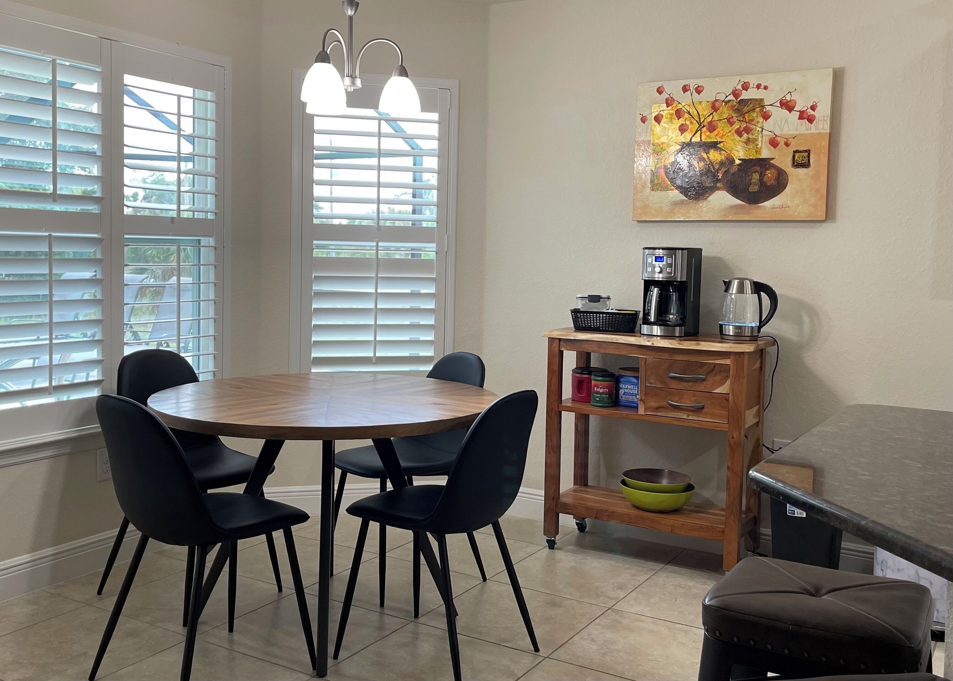 A dining area with a round table, four chairs, a coffee maker, a kettle, and a painting on the wall.
