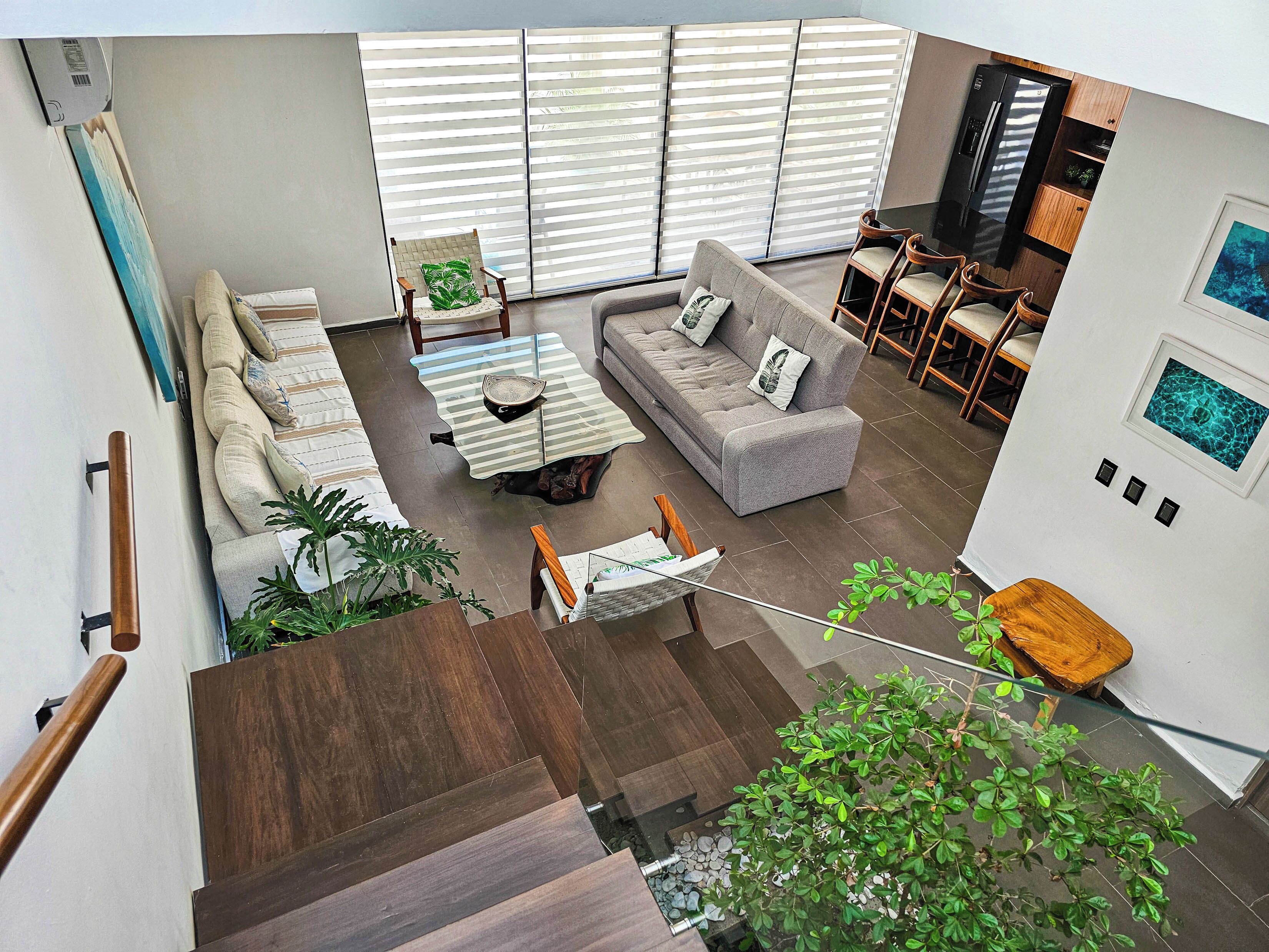 A modern living room with a sofa, coffee table, and chairs, a staircase with wooden steps, and a view of the kitchen and dining area.