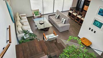A modern living room with a sofa, coffee table, and chairs, a staircase with wooden steps, and a view of the kitchen and dining area.