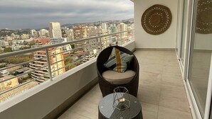 Un balcón con vistas a un paisaje urbano, una silla de mimbre con cojines y una pequeña mesa redonda con superficie de vidrio.