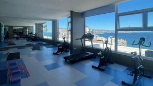 Un gimnasio con equipamiento de ejercicio y amplias ventanas con vistas a un paisaje urbano.