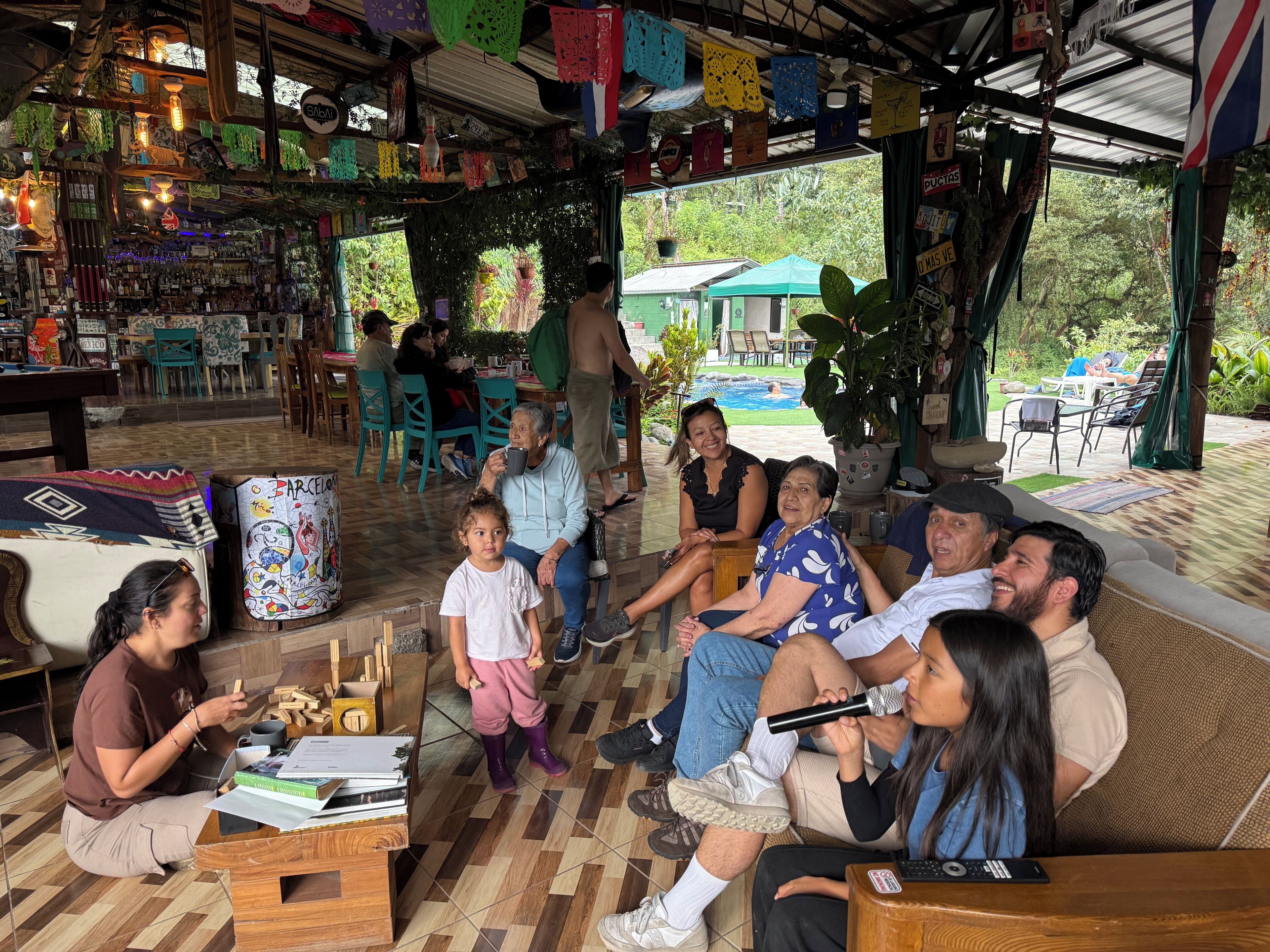Un grupo de personas sentadas en un sofá y sillas en un espacio exterior cubierto, con un bar y varios objetos en estanterías.
