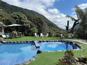 Una piscina con borde de azulejos, rodeada de rocas y césped artificial, con sillas blancas y una mesa bajo un paraguas verde, frente a un fondo de vegetación exuberante y una montaña.