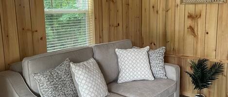 Un canapé gris avec des coussins à motifs, dans une pièce aux murs recouverts de lambris en bois, avec une grande fenêtre et une petite plante en pot.