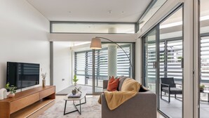 A modern living room with a flat-screen TV, a wooden console, a gray sofa, and a large window offering a view of the city. - Urban Rest Double Bay Apartments (Double Bay)