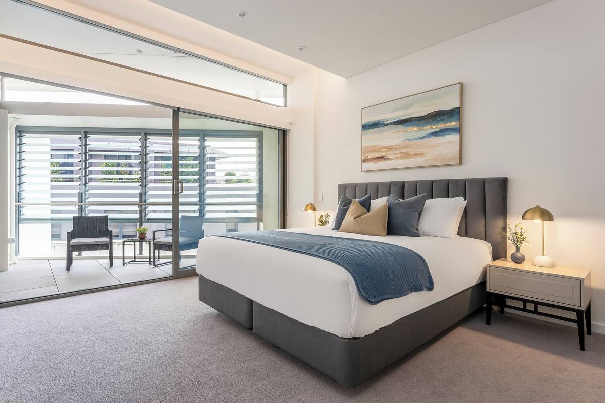 Um quarto moderno com uma cama grande, uma mesa de cabeceira com abajur e vista para uma varanda externa por meio de portas de vidro do chão ao teto.