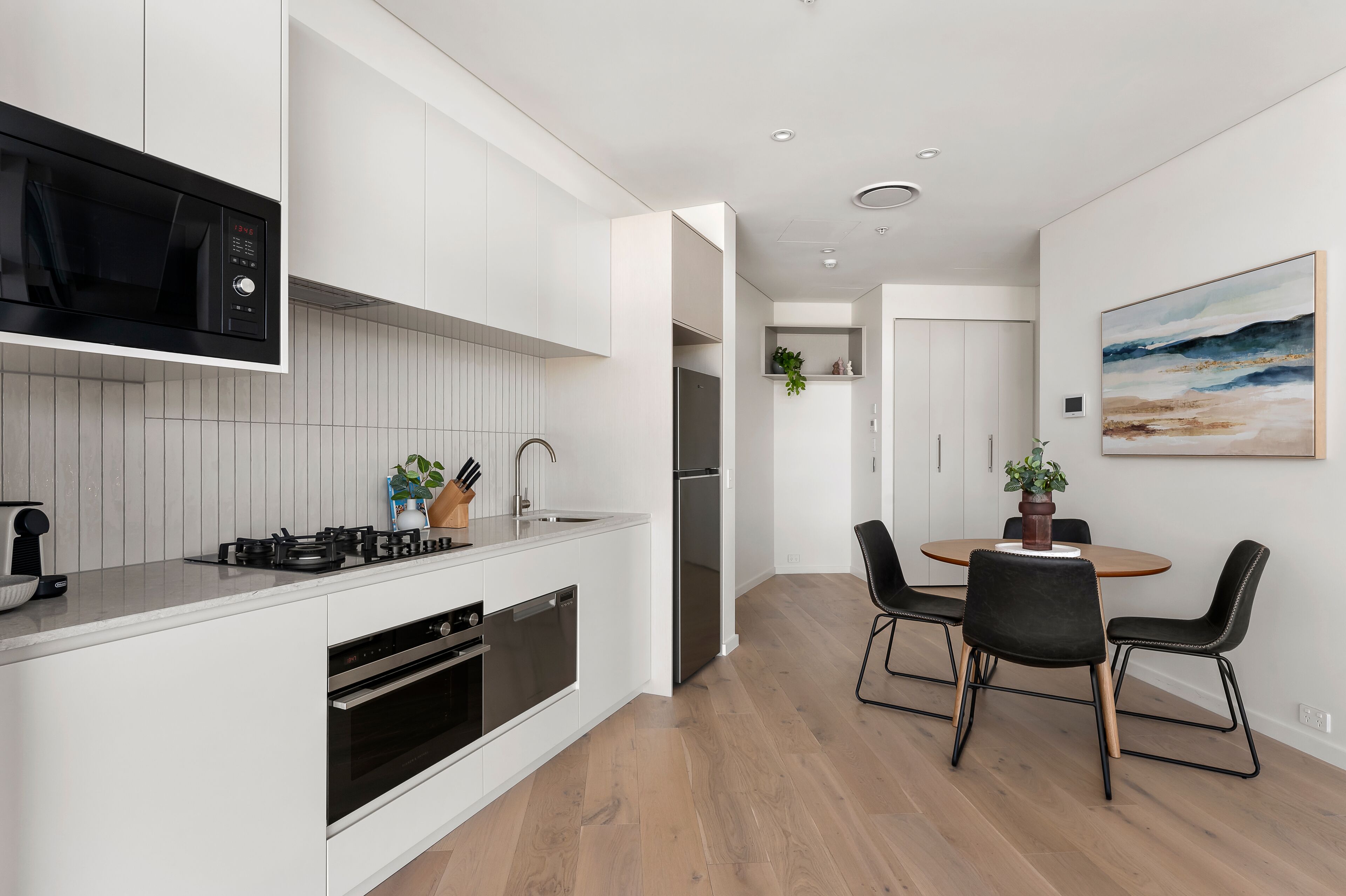 Een moderne keuken met witte kasten, een ingebouwde magnetron, gasfornuis en vaatwasser. Er is een eethoek met een ronde tafel en vier stoelen.