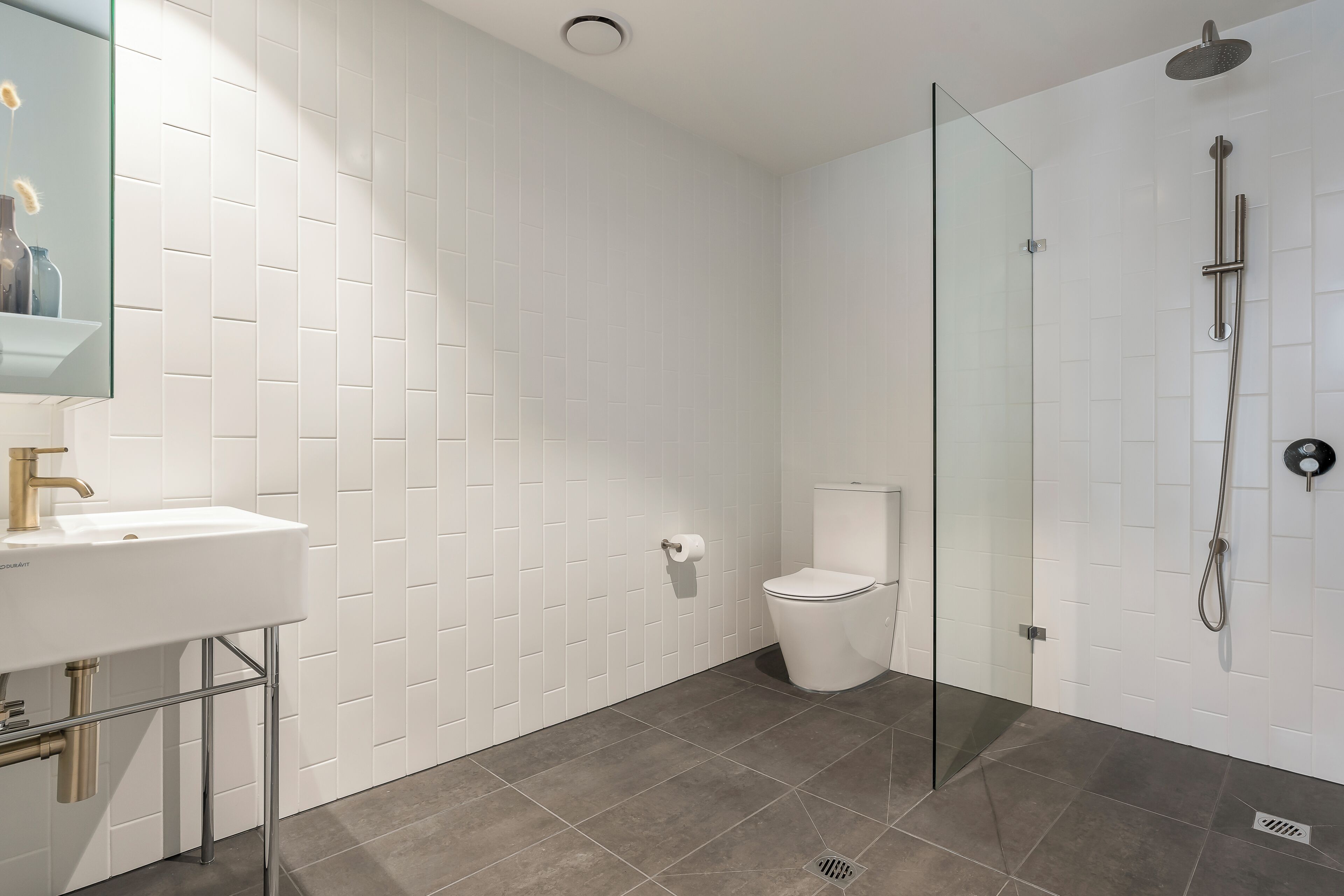 Et moderne badeværelse med hvid mosaik, en glasbrusekabine, et hvidt toilet og en vask med kromarmaturer.