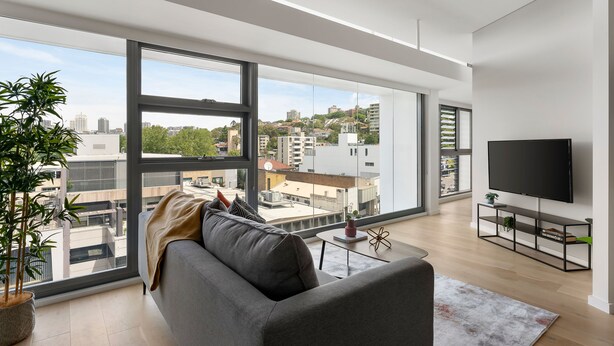 A modern living room with large windows, a grey sofa, a flat-screen TV, and a potted plant. - Urban Rest Double Bay Apartments (Double Bay)