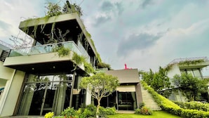 A modern multi-story house with a glass facade, a flat roof, and a small garden area with a tree and shrubs.