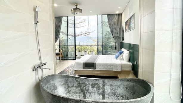 A modern bathroom with a large freestanding bathtub, a glass-enclosed shower area, and a bed with a view of the outdoors.