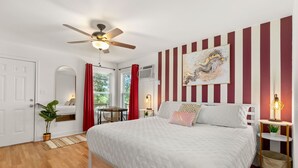 Un dormitorio con una cama, ventilador de techo, ventana con cortinas rojas, aire acondicionado de pared y un cuadro.