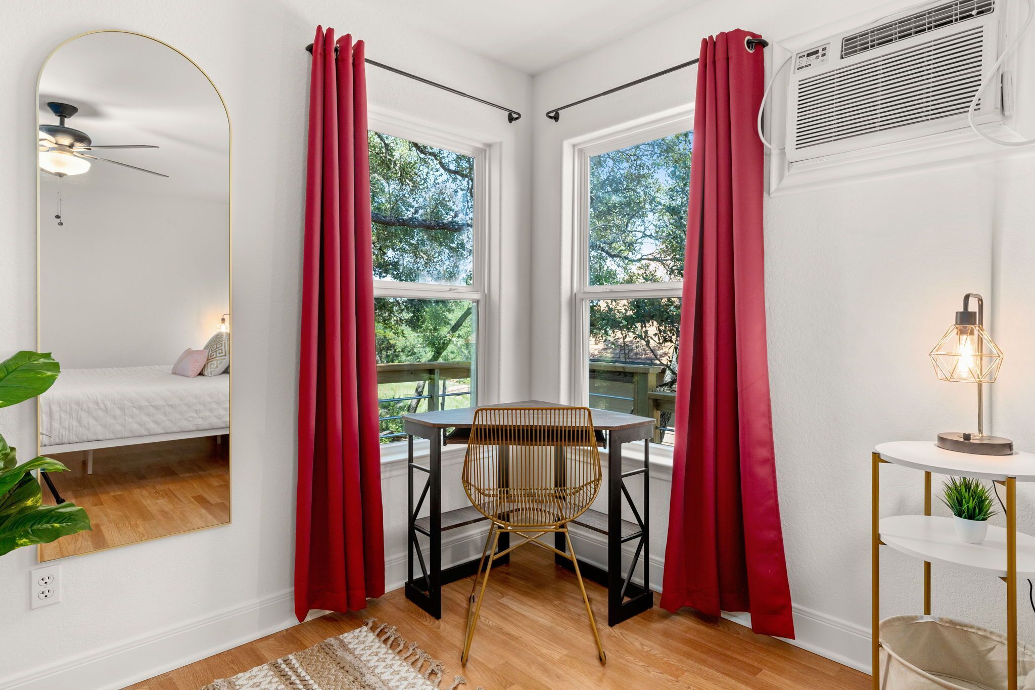 A corner room with a bed, a table with a chair, a mirror, and a window with red curtains.