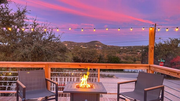 Una terraza con un hoyo para el fuego, dos sillas y luces colgantes, sobre un fondo de atardecer.