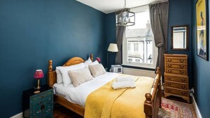 A bedroom with a bed, a nightstand, a dresser, a window with curtains, and a view of buildings outside.