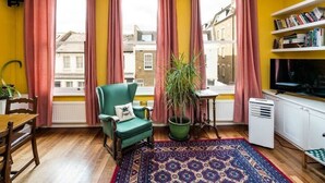 A cozy living room with a green armchair, a potted plant, a patterned rug, and a bookshelf.