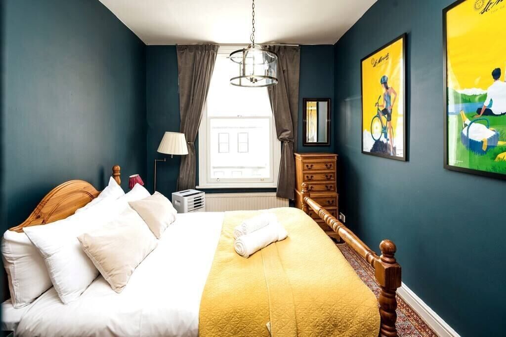 A bedroom with a bed, a wooden headboard, a yellow bedspread, a white lamp, a dresser, and a window with curtains.