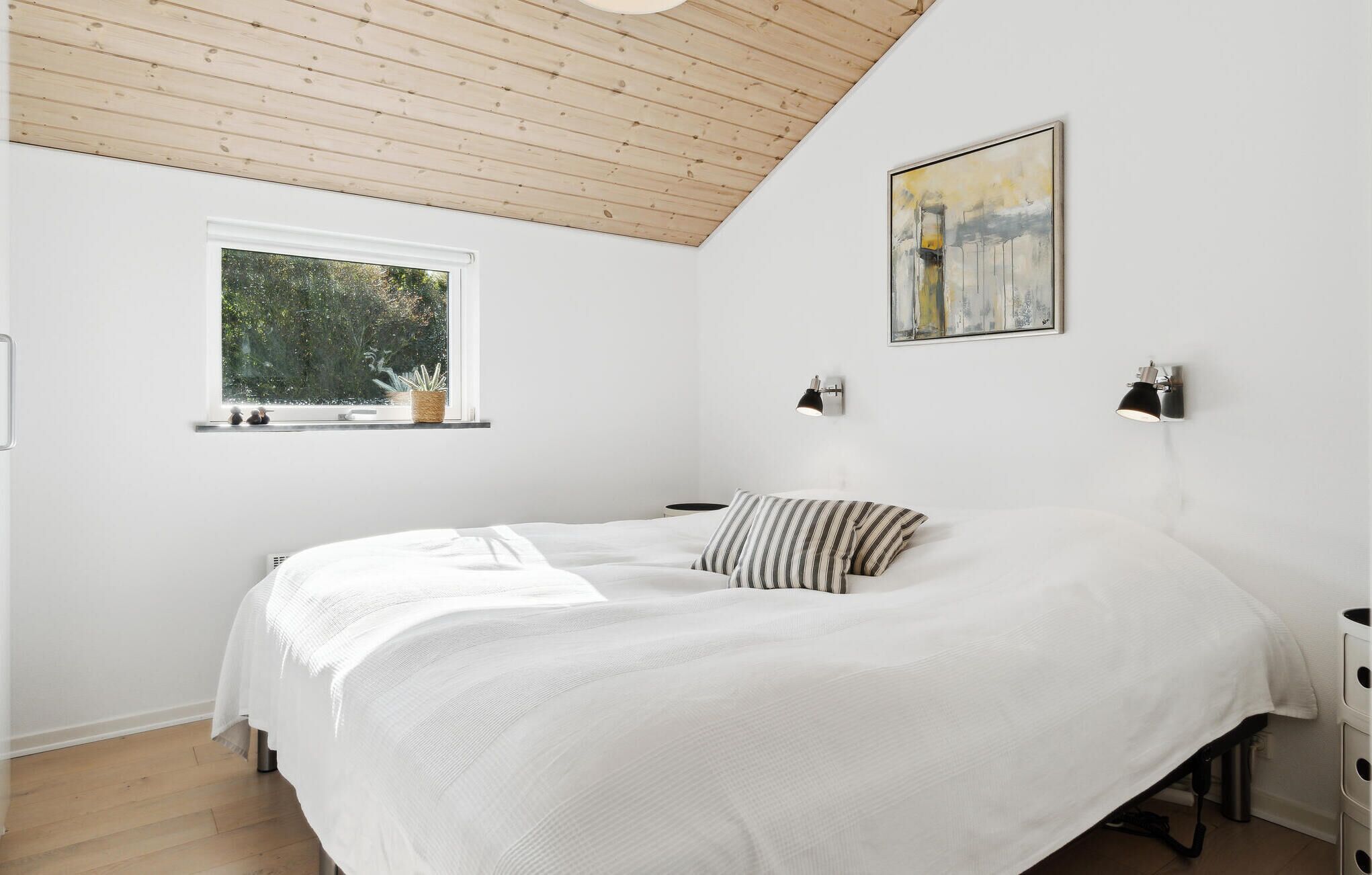 Un dormitorio con techo de madera, un ventanal con vistas a la vegetación, una cama con ropa blanca, dos lámparas de noche y un cuadro en la pared.