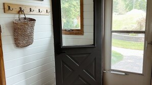 A black door with a window, a woven basket hanging on a wall-mounted rack, and a doormat with a message.