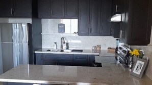 A modern kitchen with dark cabinets, a granite countertop, a stainless steel refrigerator, a sink, and two white bar stools. - THE AQUEDUCTS OF NEGRIL
"Seaview Paradise" (Negril)