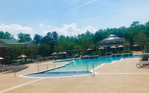 Une piscine entourée d’arbres et d’un bâtiment à colonnes, avec des chaises longues et des parasols à proximité.