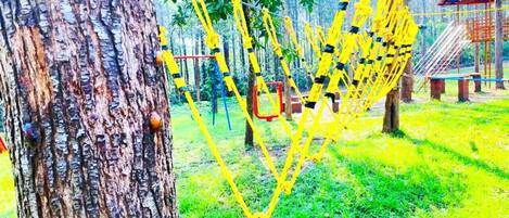 A rope bridge in a park with trees and a playground structure in the background.