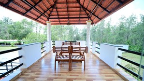 A covered outdoor seating area with wooden benches and a tiled floor, surrounded by white railings and columns, overlooking a forested area.