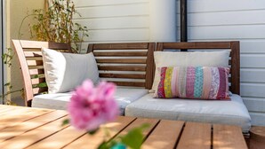 Un banco de madera con cojines blancos y un cojín de retazos coloridos, una mesa de madera y una flor rosa en un jarrón azul.