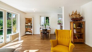 Un salón luminoso con un sillón amarillo, estanterías de madera, una mesa de comedor con sillas y un ventanal grande.