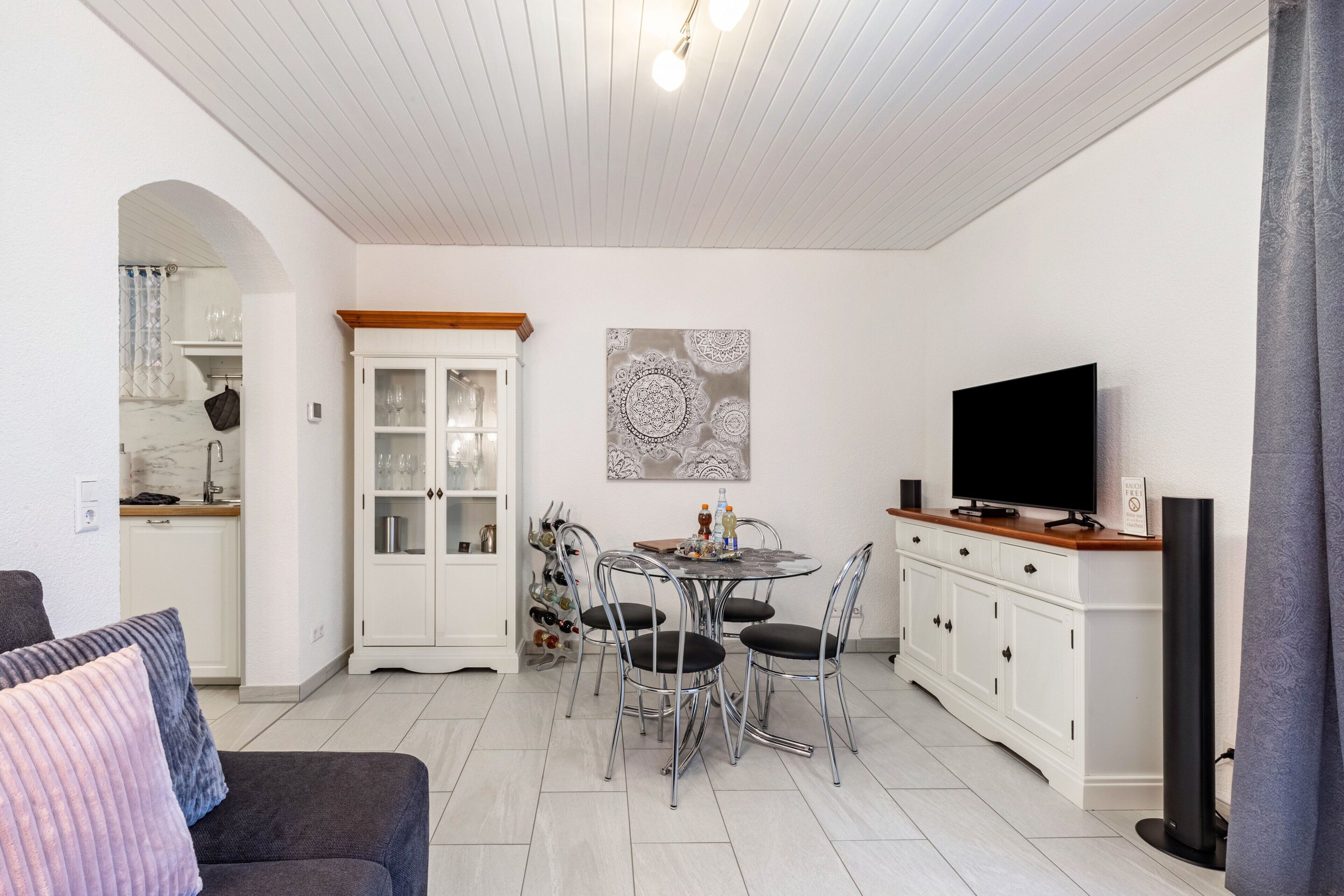 A dining area with a round table, chairs, and a television. There is a kitchen area with cabinets and a countertop.