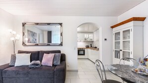 Ein modernes Wohnzimmer mit einer Couch, einem Esstisch mit Stühlen, einer Küche im Hintergrund und einem großen Spiegel an der Wand.