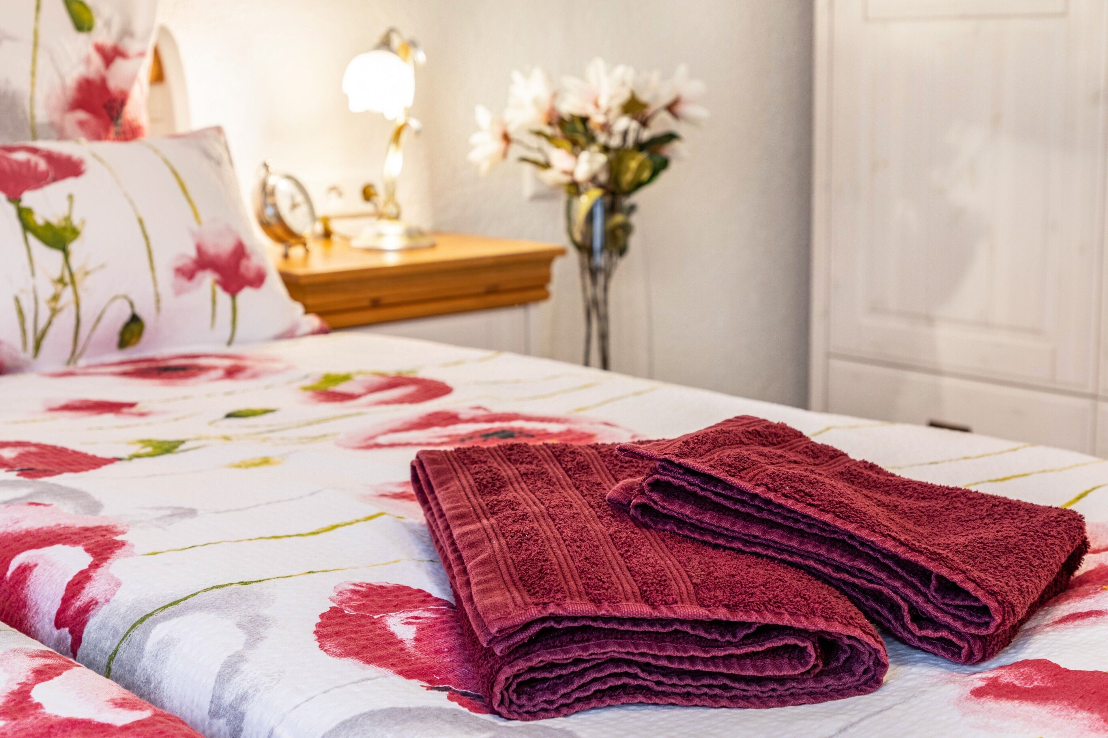 Un lit bien fait, recouvert d’une couvre-lit à motifs floraux, une pile de serviettes pliées, une lampe de chevet et un vase rempli de fleurs.