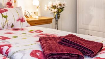 Un lit bien fait, recouvert d’une couvre-lit à motifs floraux, une pile de serviettes pliées, une lampe de chevet et un vase rempli de fleurs.