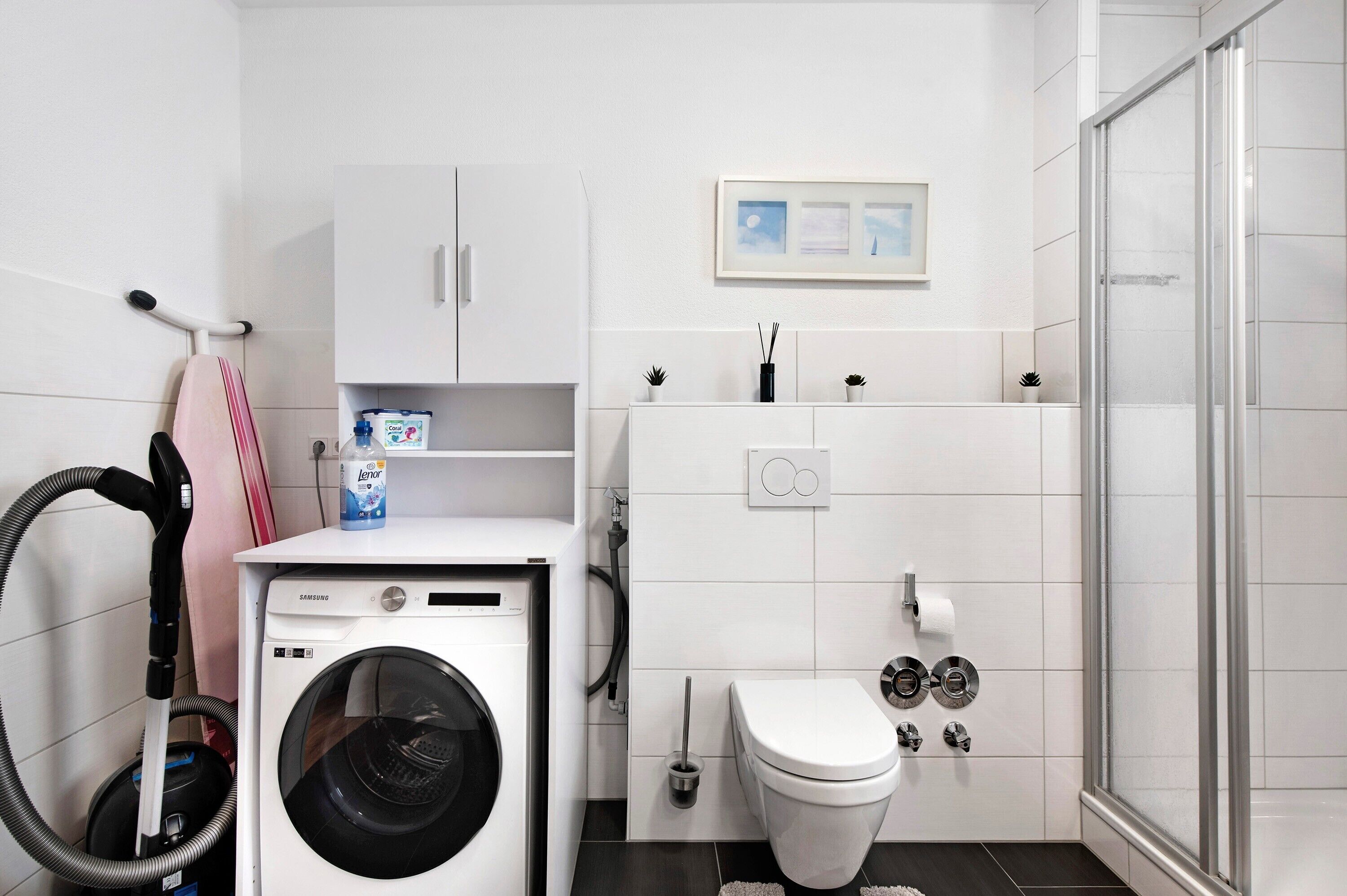 Ein kleines Hotelbad mit Waschmaschine, Toilette, Dusche und einer Ablage für Pflegeprodukte.